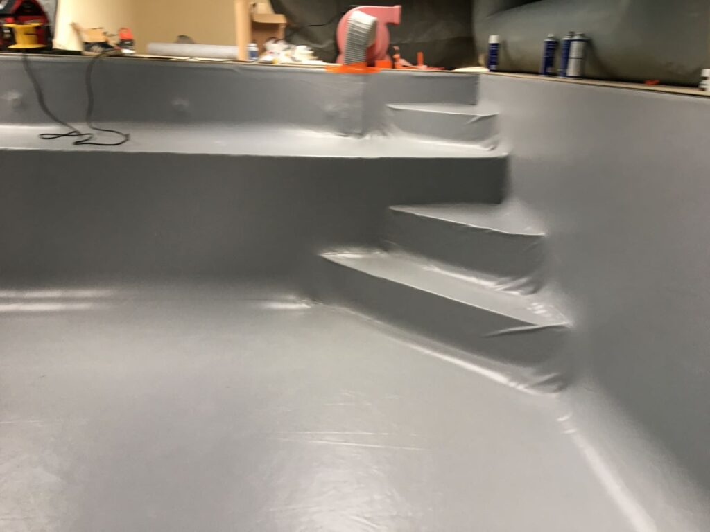 Intérieur d'une piscine en cours de rénovation, revêtement en vinyle gris neuf, lisse et tendu recouvrant les marches d'accès.