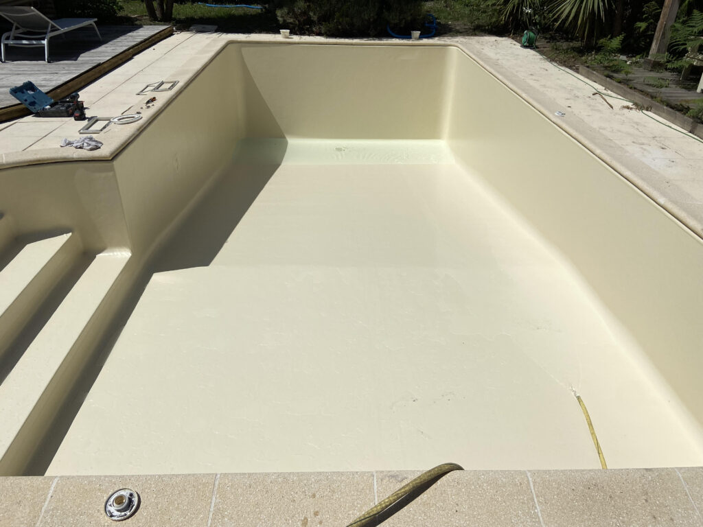 Piscine creusée vide, recouverte d'un revêtement neuf de couleur crème, avec des équipements de rénovation sur la margelle.