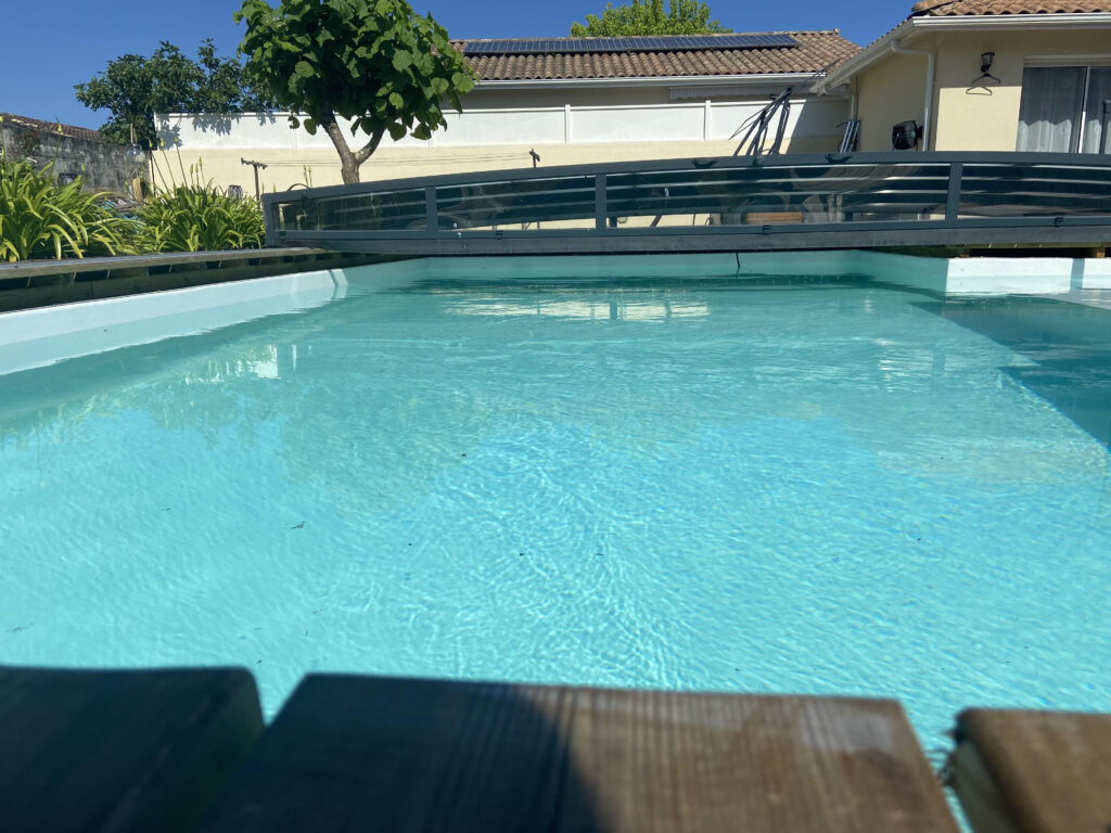 Vue en plongée d'une piscine aux eaux turquoise, avec une couverture de piscine rétractable sombre et des plantes à fleurs près d'une maison avec panneaux solaires.