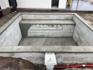 Piscine creusée en béton brute, avec des conduits et une pièce de skimmer visibles, en cours de construction à côté d'une terrasse en bois.