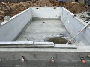 Piscine en construction : structure en béton brut fraîchement crépie, avec tas de terre à l'arrière et équipement de maçonnerie.
