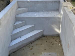Escalier en béton brut pour une piscine non remplie, avec une finition projetée et des marches inclinées, jonché de feuilles mortes.