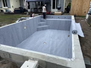 Installation d'un revêtement de fond de piscine en feutre gris (LIPUSSE 10 GRIS 2025) dans une coque en béton brute, un ouvrier est debout sur les marches.