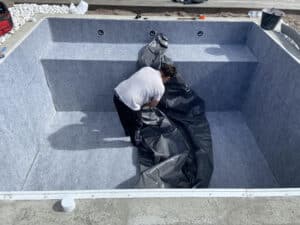 Installation liner piscine : ouvrier déploie un grand revêtement gris foncé dans un bassin préparé avec un feutre gris marqué "LIPLISSE 10 GRIS 2024".