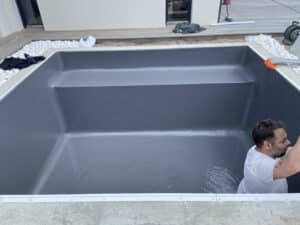 Installation d'un liner de piscine gris foncé, un homme en t-shirt blanc travaille dans l'eau peu profonde autour du bord.