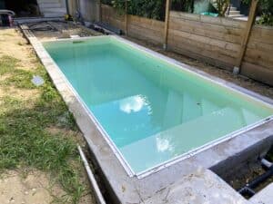 Piscine coque hors-sol ou semi-enterrée en cours d'installation, remplie d'eau claire reflétant le ciel, avec une margelle en béton et un chantier autour.
