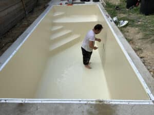 Un ouvrier torse nu travaille pieds nus dans une piscine creusée vide, ajustant une pièce avec un tournevis près d'une bonde de refoulement.