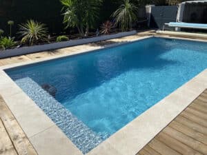 Belle piscine rectangulaire aux eaux bleues éclatantes, bordée de margelles claires et d'une terrasse en bois sous le soleil.
