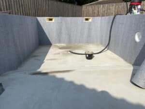 Construction de piscine : revêtement en feutre gris posé sur la dalle de fond, avec des ouvertures pour les skimmers et un seau de chantier.