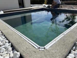 Construction d'une piscine enterrée avec margelle texturée, système de fixation blanc, et galets blancs décoratifs, un ouvrier travaille à proximité.