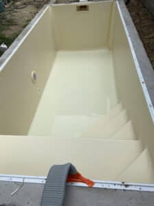Vue intérieure d'un nouveau bassin de piscine coque couleur crème, avec escalier intégré et margelle temporaire. Tuyau gris apparent.
