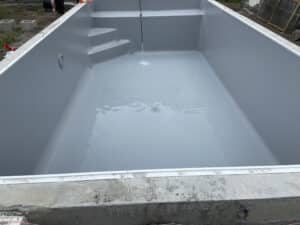 Piscine coque gris clair neuve se remplissant d'eau, avec escalier intégré et fixation du liner visible sur le pourtour en béton.