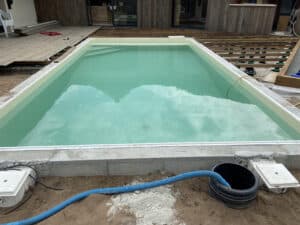 Piscine coque partiellement enterrée, remplie d'eau turquoise, chantier de terrasse bois en cours avec lambourdes visibles.