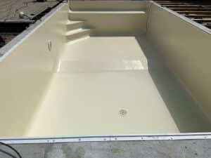 Coque de piscine en fibre de verre beige clair, neuve, partiellement remplie d'eau, avec escaliers intégrés et bonde de fond visible, en cours d'installation.