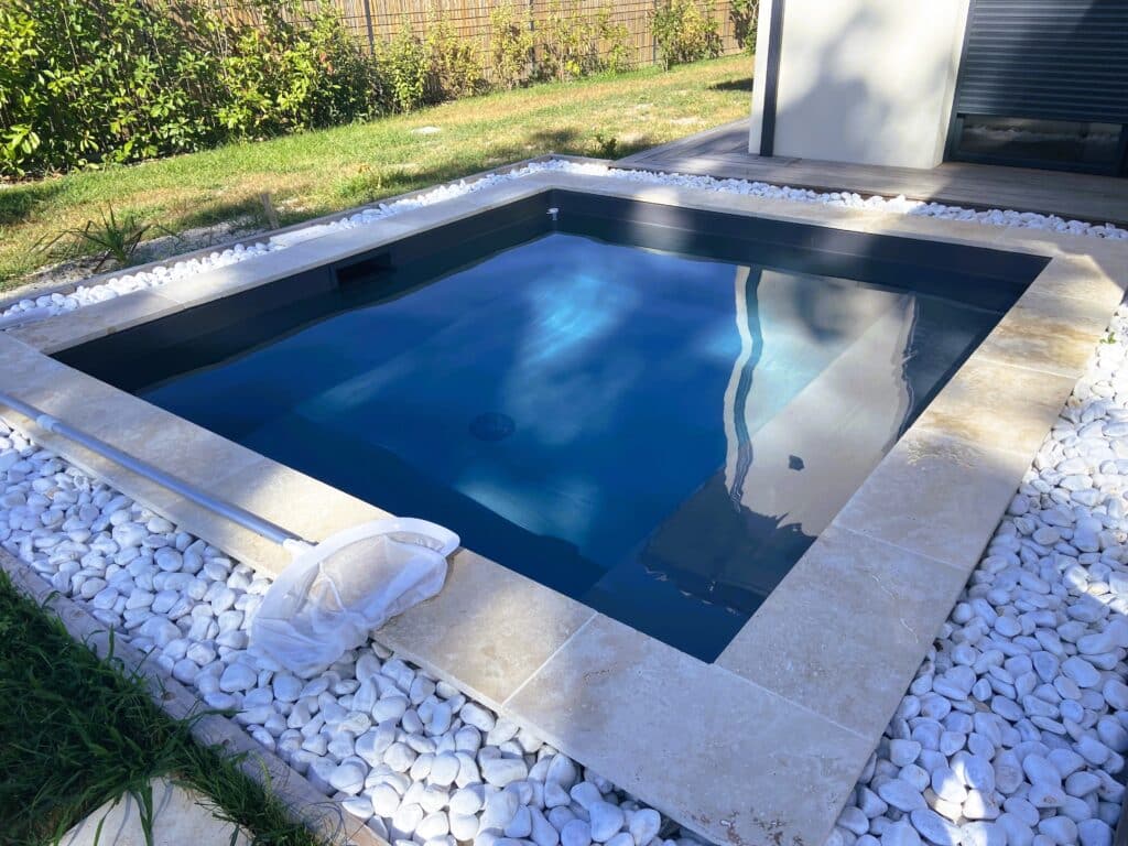 Petite piscine rectangulaire moderne avec eau bleue, margelle en pierre claire et galets blancs, près d'un mur et d'un espace en bois.