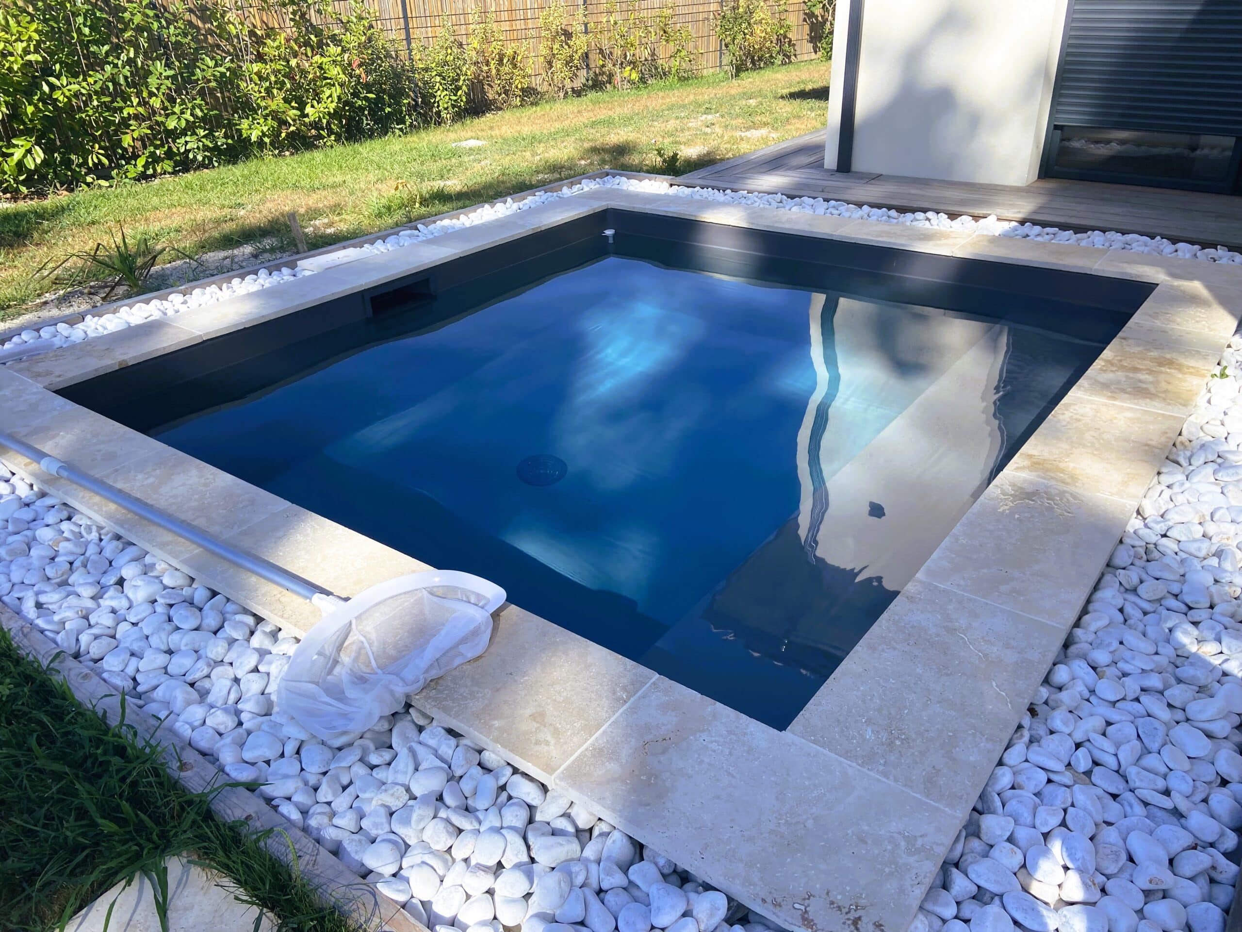 Petite piscine rectangulaire moderne avec eau bleue, margelle en pierre claire et galets blancs, près d'un mur et d'un espace en bois.