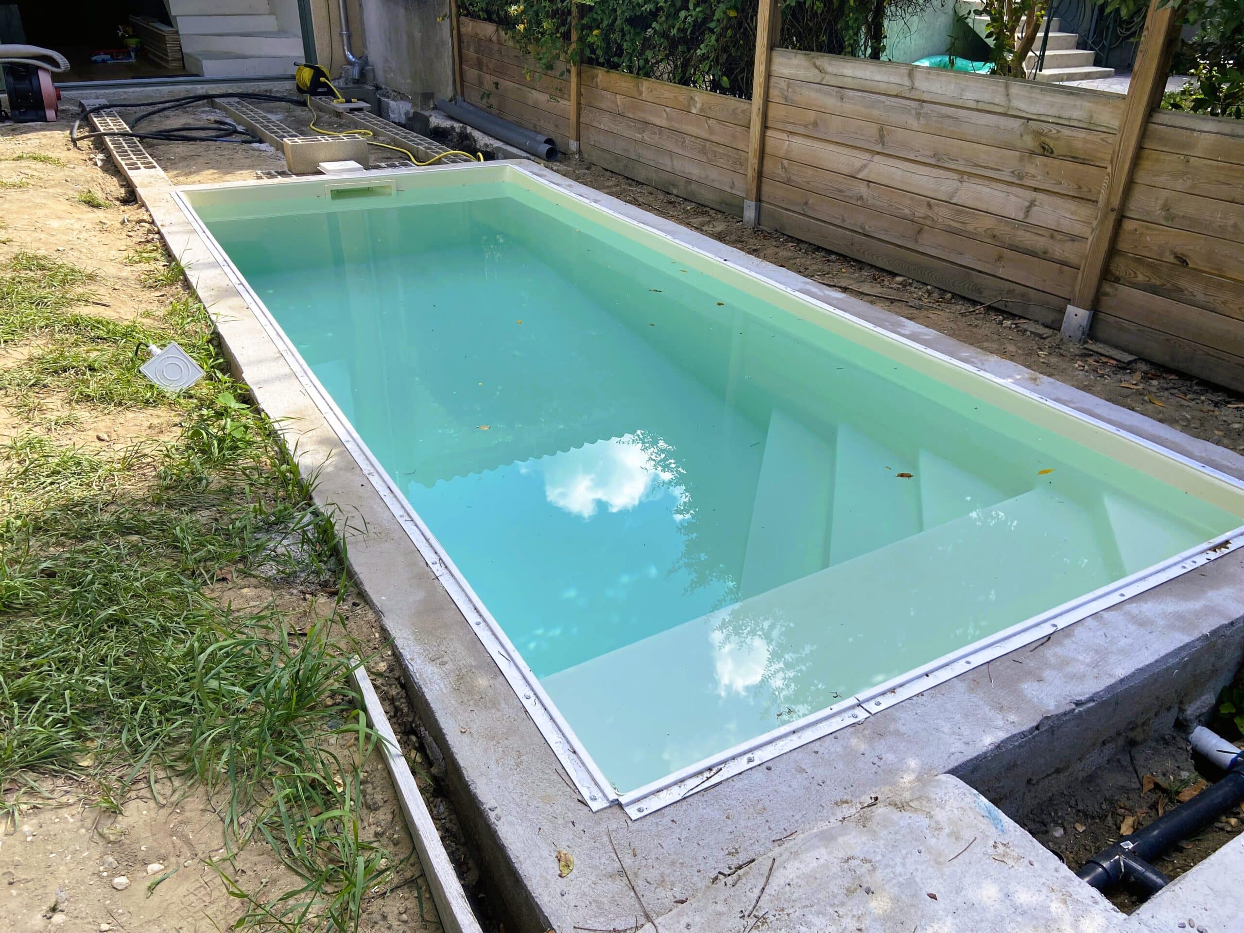 Installation piscine coque chantier et clôture bois Piscine hors-sol en cours d'installation, remplie d'eau turquoise, bordée de béton brut et d'une clôture en bois, chantier environnant.