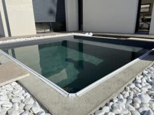 Petite piscine carrée moderne remplie d'eau vert foncé, bordée de béton brut et de galets blancs, devant une façade claire avec volet roulant.