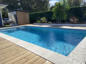 Piscine rectangulaire aux eaux bleues claires, bordée d'une terrasse en bois et de margelles en pierre, avec un abri en bois et une haie en arrière-plan.