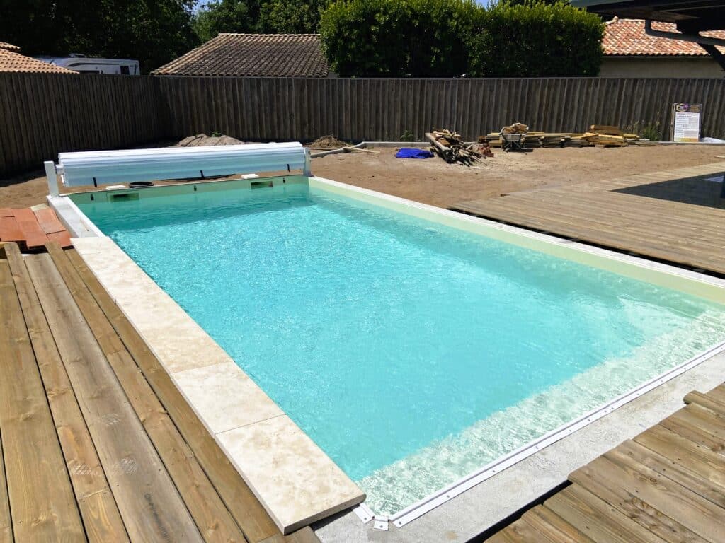 Piscine hors-sol moderne remplie d'eau turquoise, bordée d'une margelle en pierre claire et d'une terrasse en bois en cours d'installation. Un enrouleur de couverture est visible.