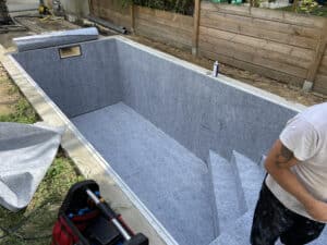 Installation d'un feutre de protection gris 'LIPUSSE 10 GRIS 2024' à l'intérieur d'une coque de piscine vide, un ouvrier est visible.