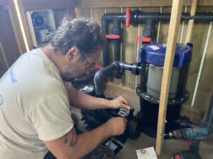 Homme avec barbe et lunettes, torse portant un t-shirt "sunrise", travaille sur le câblage électrique d'une pompe de piscine à côté d'un filtre et de vannes rouges.