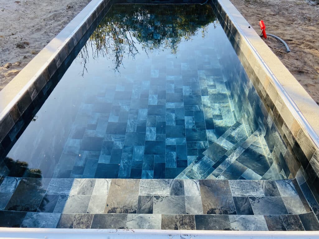 Carrelage piscine ardoise bleue : design moderne et élégant. Piscine en construction avec carrelage ardoise bleu profond. L'eau claire reflète les arbres. Margelle et sable.