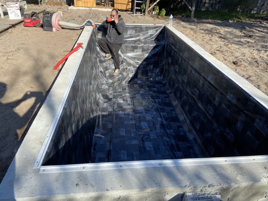 Pose du liner de piscine vinyle à motif ardoise. Un installateur sécurise le revêtement foncé près de la lèvre en béton, avec un ventilateur prêt.